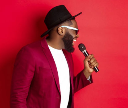 A black man wearing a hat and a jacket singing into a microphone against a red background.