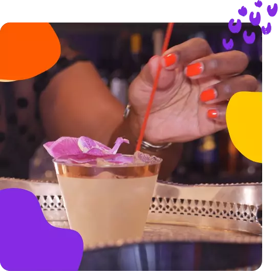 A woman is holding a cocktail in front of a colorful background.